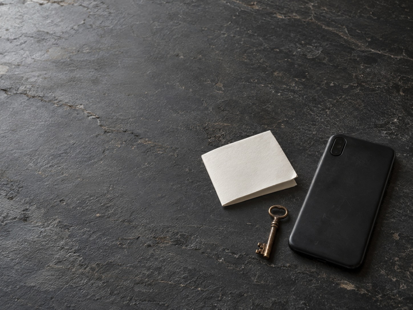 A face-down smartphone next to a small brass key and a folded blank sheet of paper on a slate countertop.