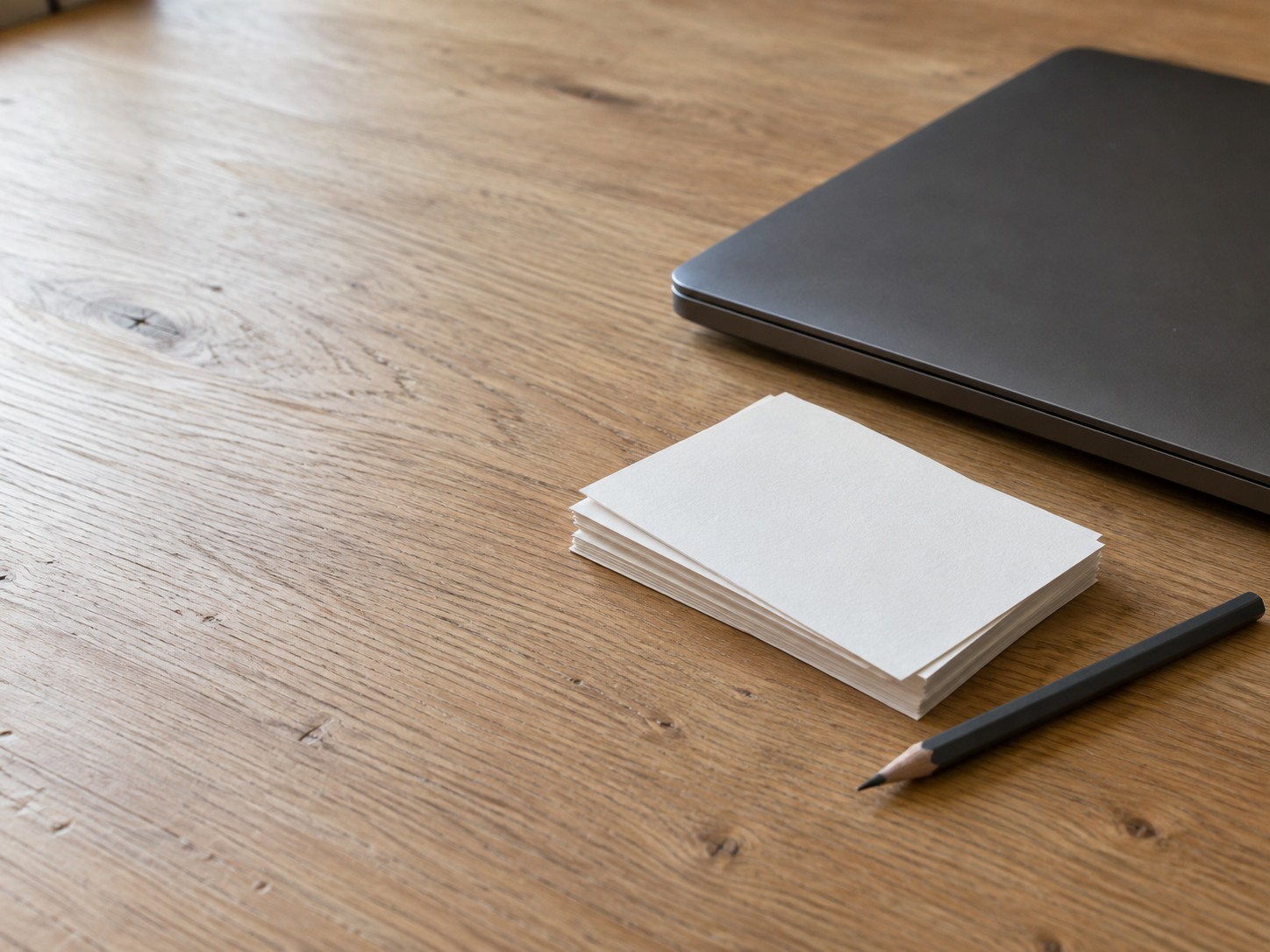 A closed laptop beside a short stack of blank index cards and a pencil on a light oak desk.