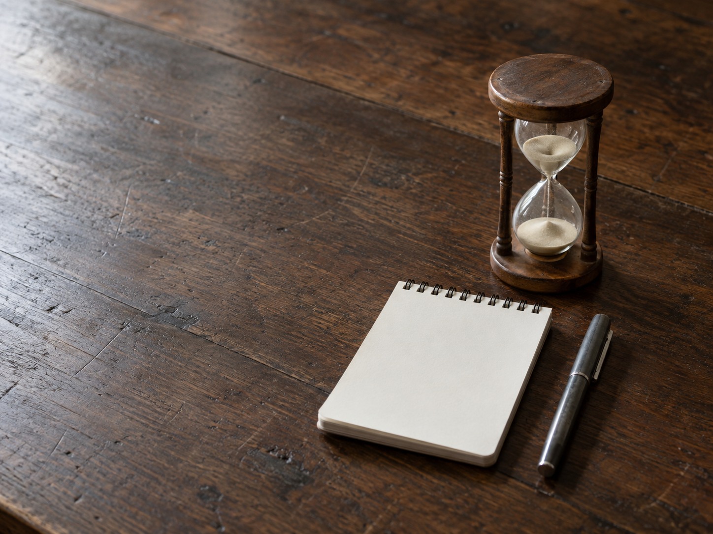 A small sand hourglass, a blank notepad and a capped pen arranged on a walnut table in soft morning light.