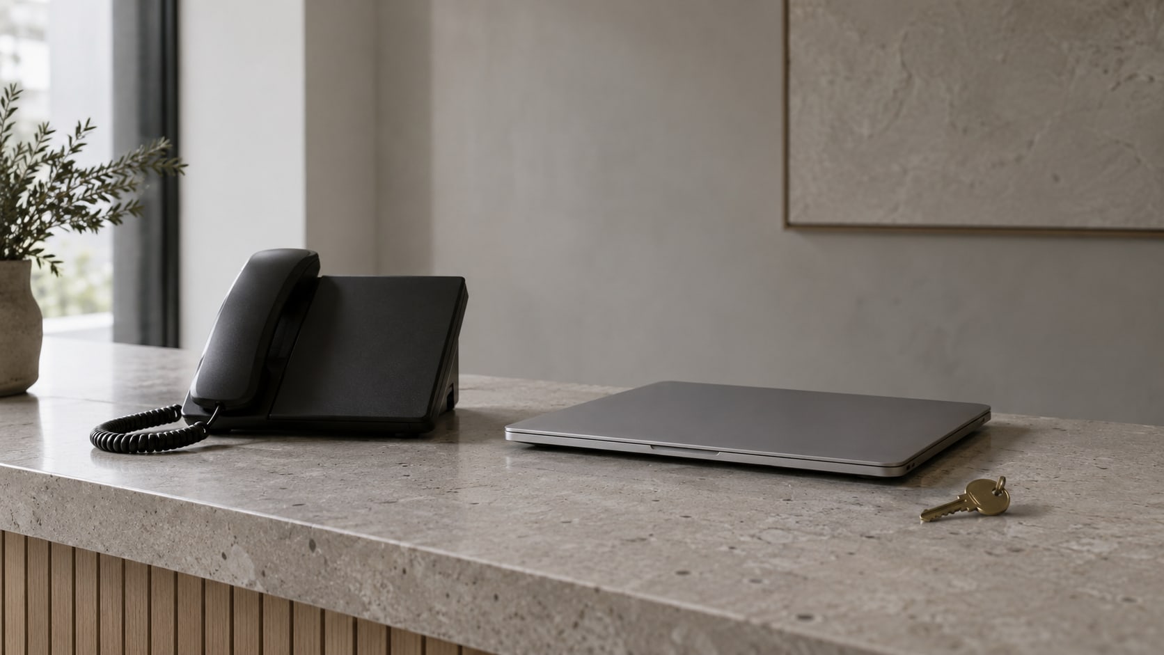 A desk phone and a closed laptop side by side on a quiet reception counter, with a small brass key beside them.