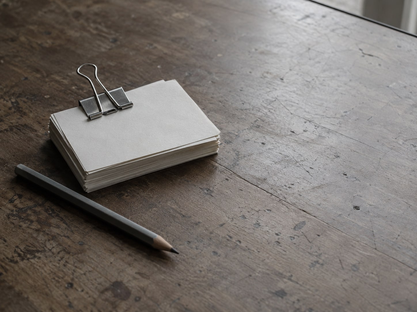 A stack of blank index cards held with a metal clip beside a pencil on a worn counter.