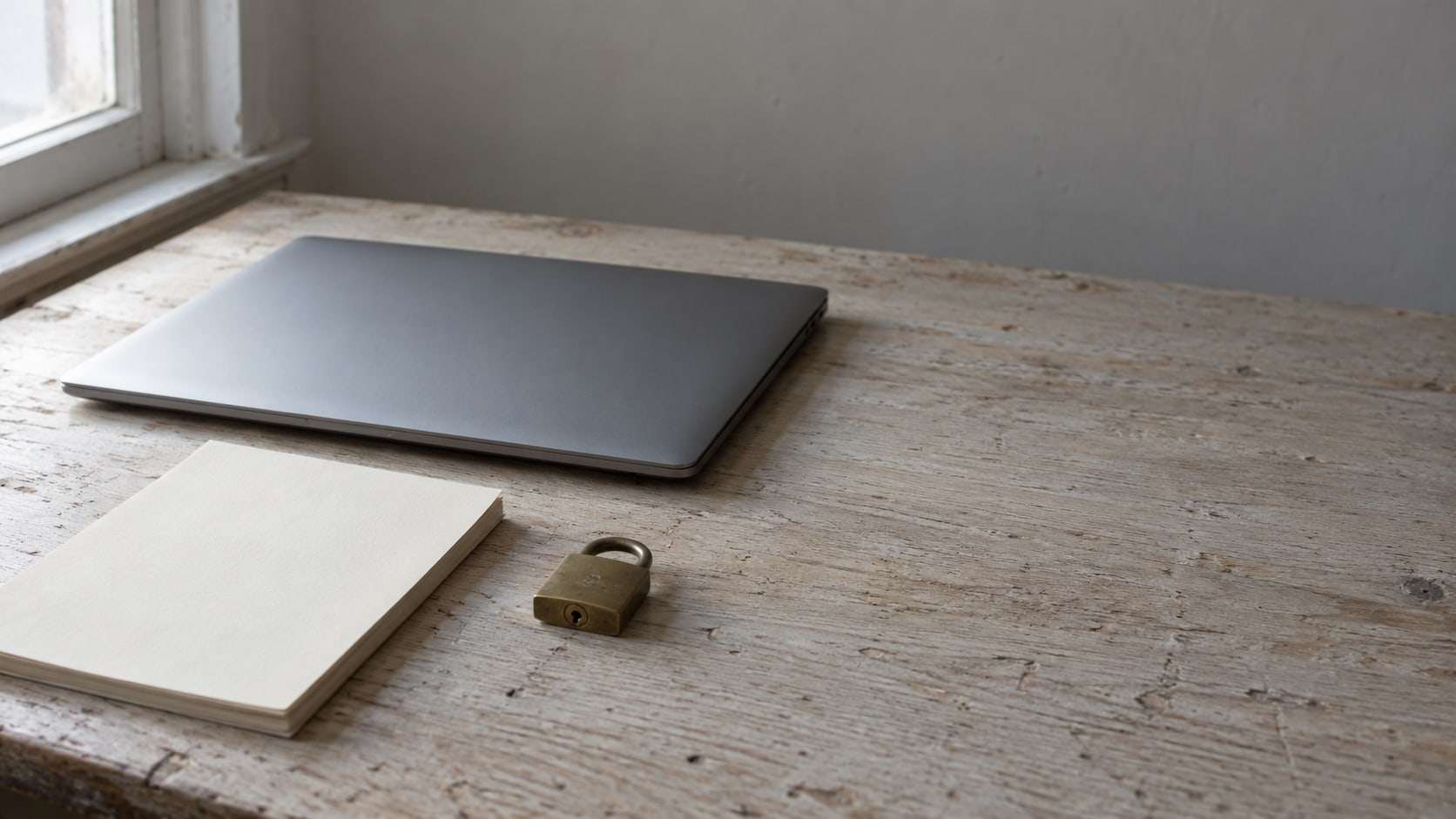 A closed laptop, a blank notepad and a small brass padlock arranged on a pale wooden desk.