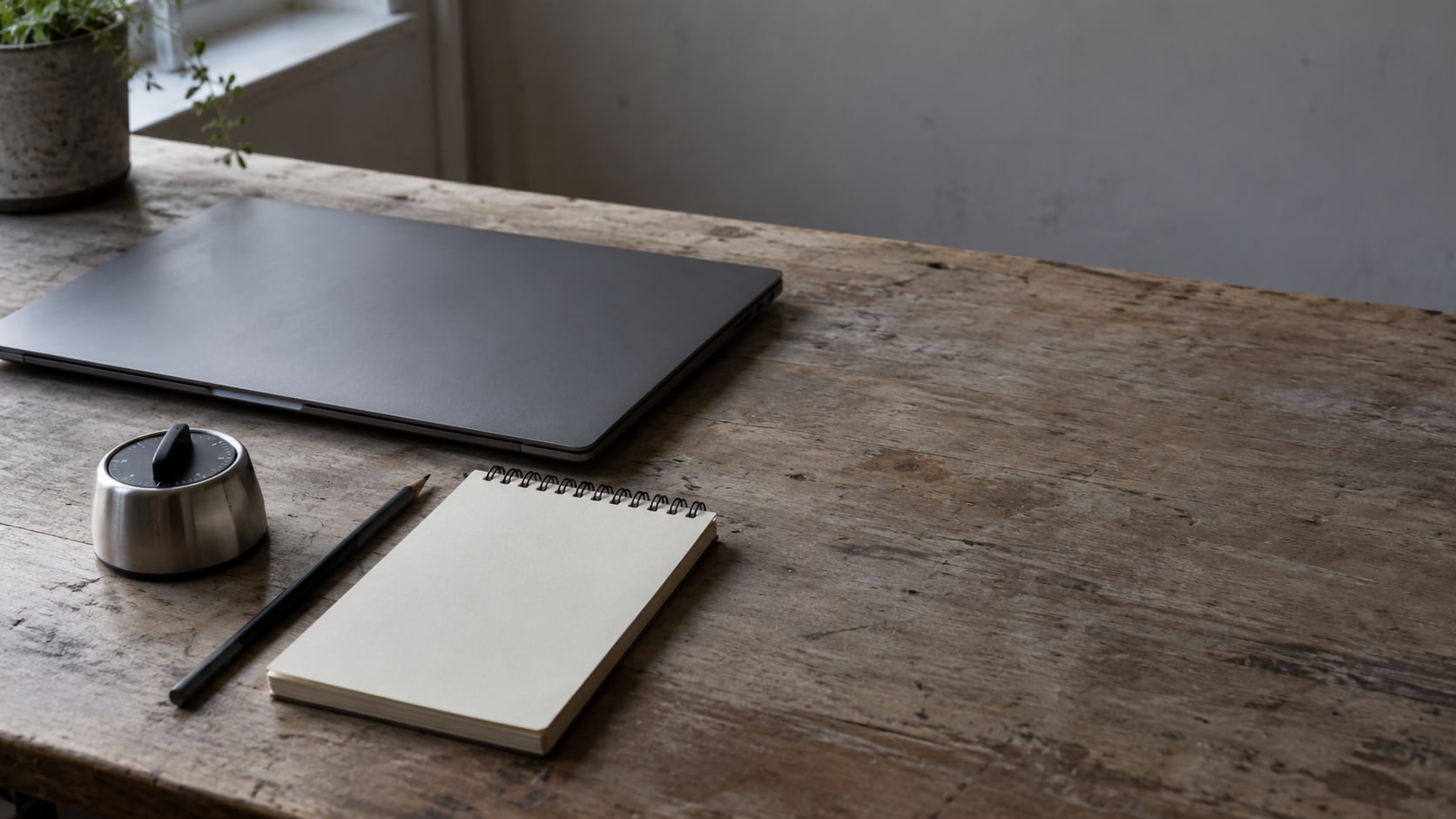 Una laptop cerrada junto a un pequeño temporizador de cocina y un bloc de notas en blanco sobre un escritorio de madera gastada.