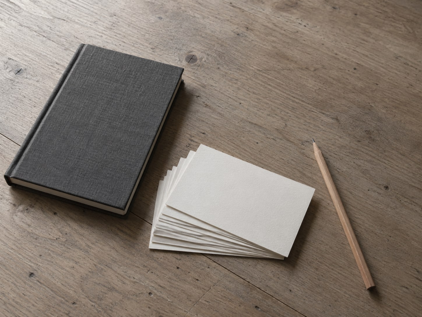 A small stack of blank index cards fanned out on a wooden desk beside a closed notebook and a sharpened pencil.
