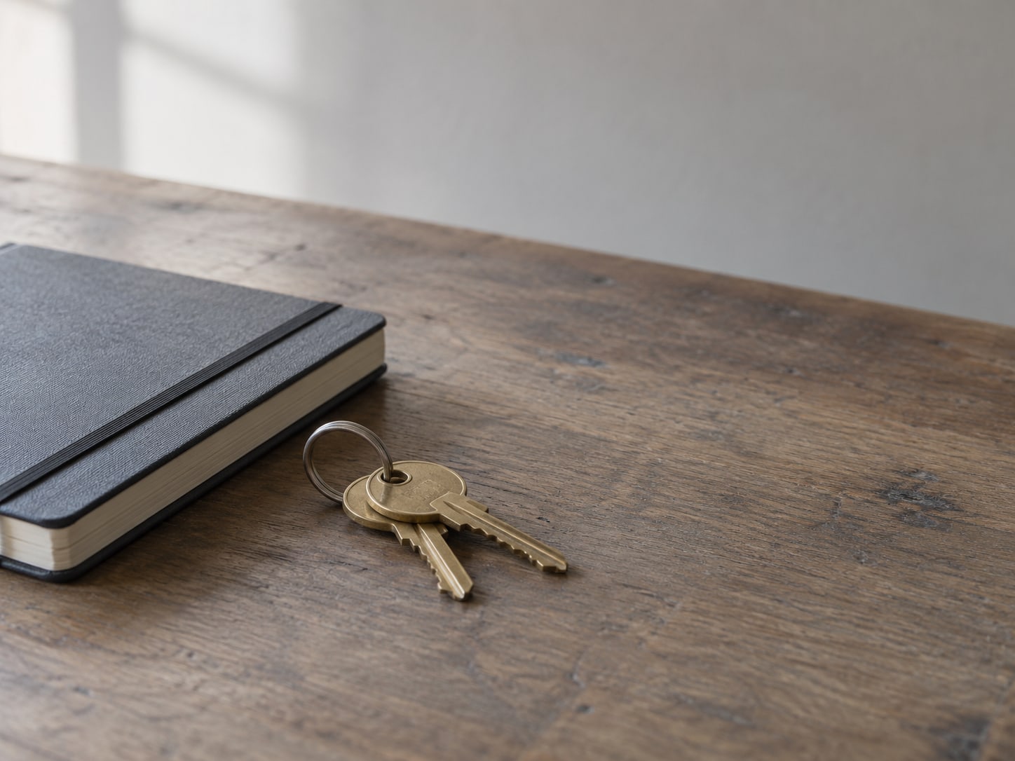A key ring with two brass keys lying beside a closed notebook on a wooden desk.