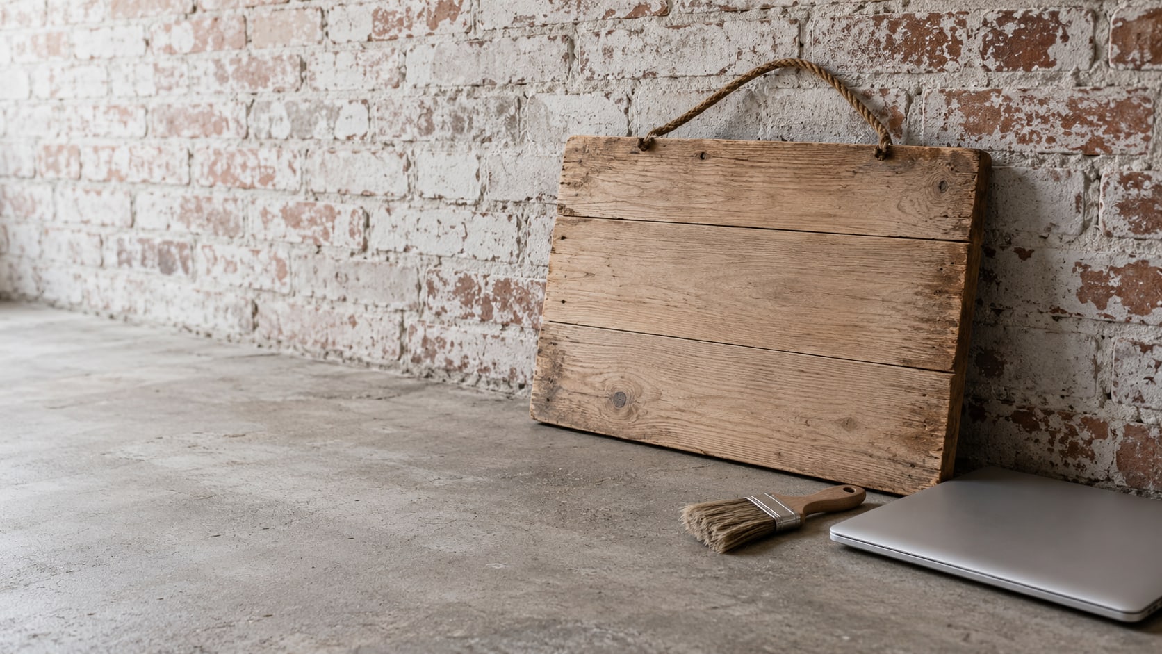 A blank wooden shop sign leaning against a brick wall beside a small paintbrush and a closed laptop.