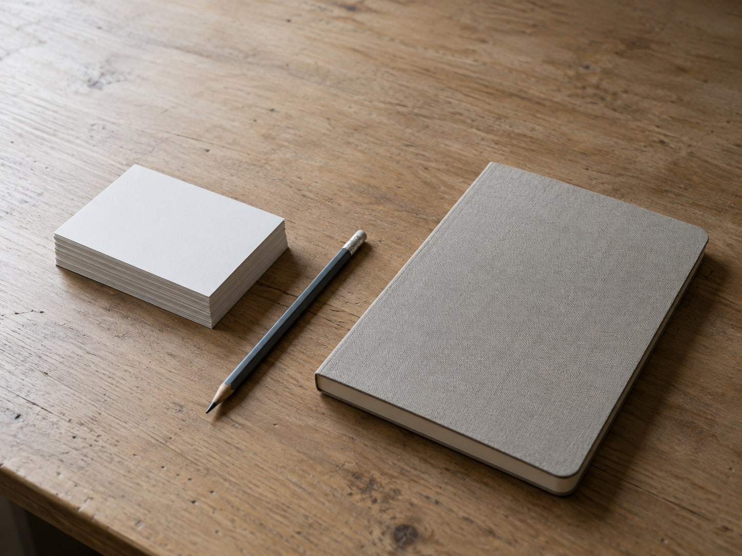 A stack of blank index cards and a pencil lined up beside a closed notebook on a clean wooden desk.