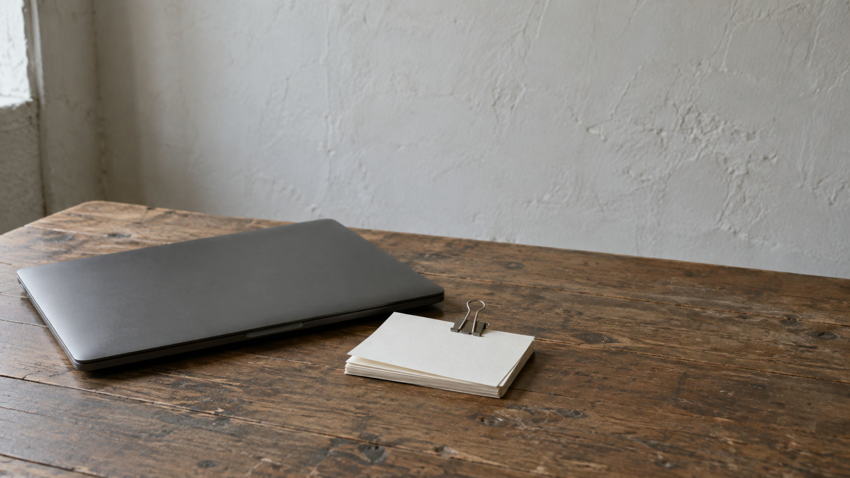 A closed laptop beside a small stack of blank index cards held with a binder clip on a wooden table.