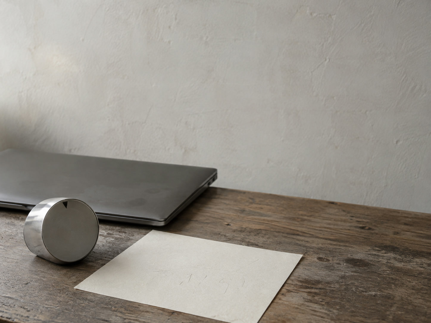 A kitchen timer next to a single sheet of blank paper with faint pencil ticks and a closed laptop on a desk.