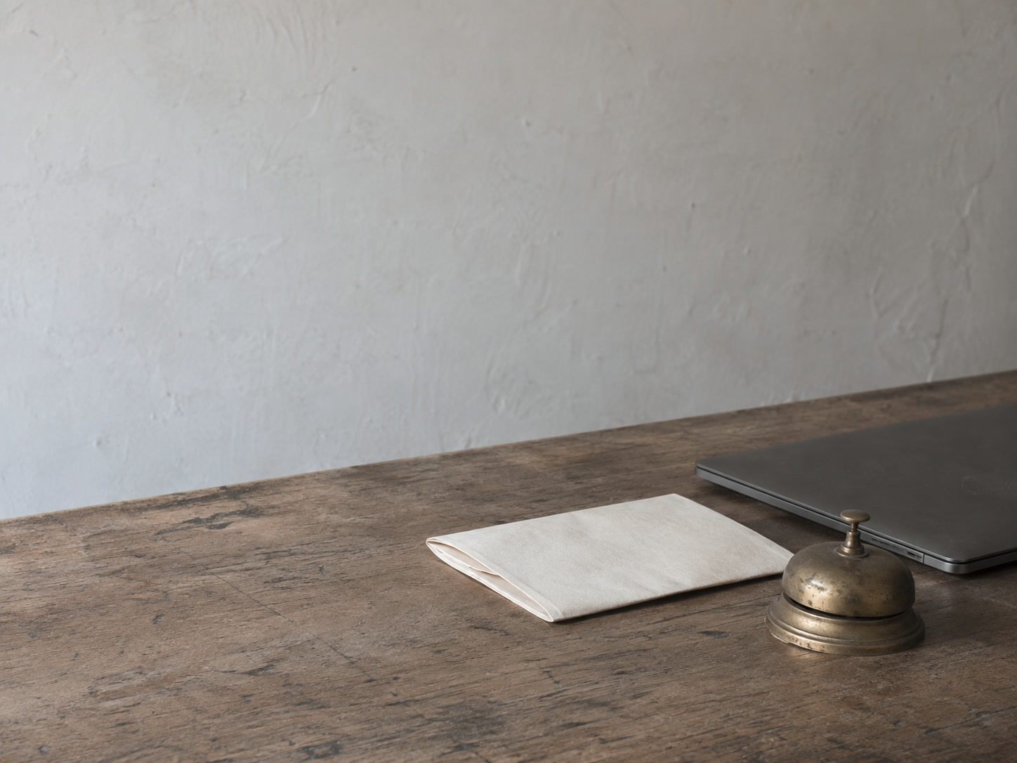 A small brass desk bell beside a folded blank letter and a closed laptop on a plain wooden counter.
