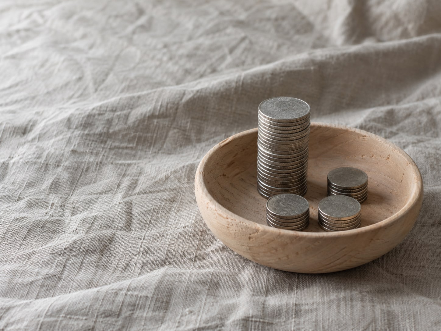 A small wooden bowl holding a single tall stack of coins beside three much smaller stacks on a linen cloth.