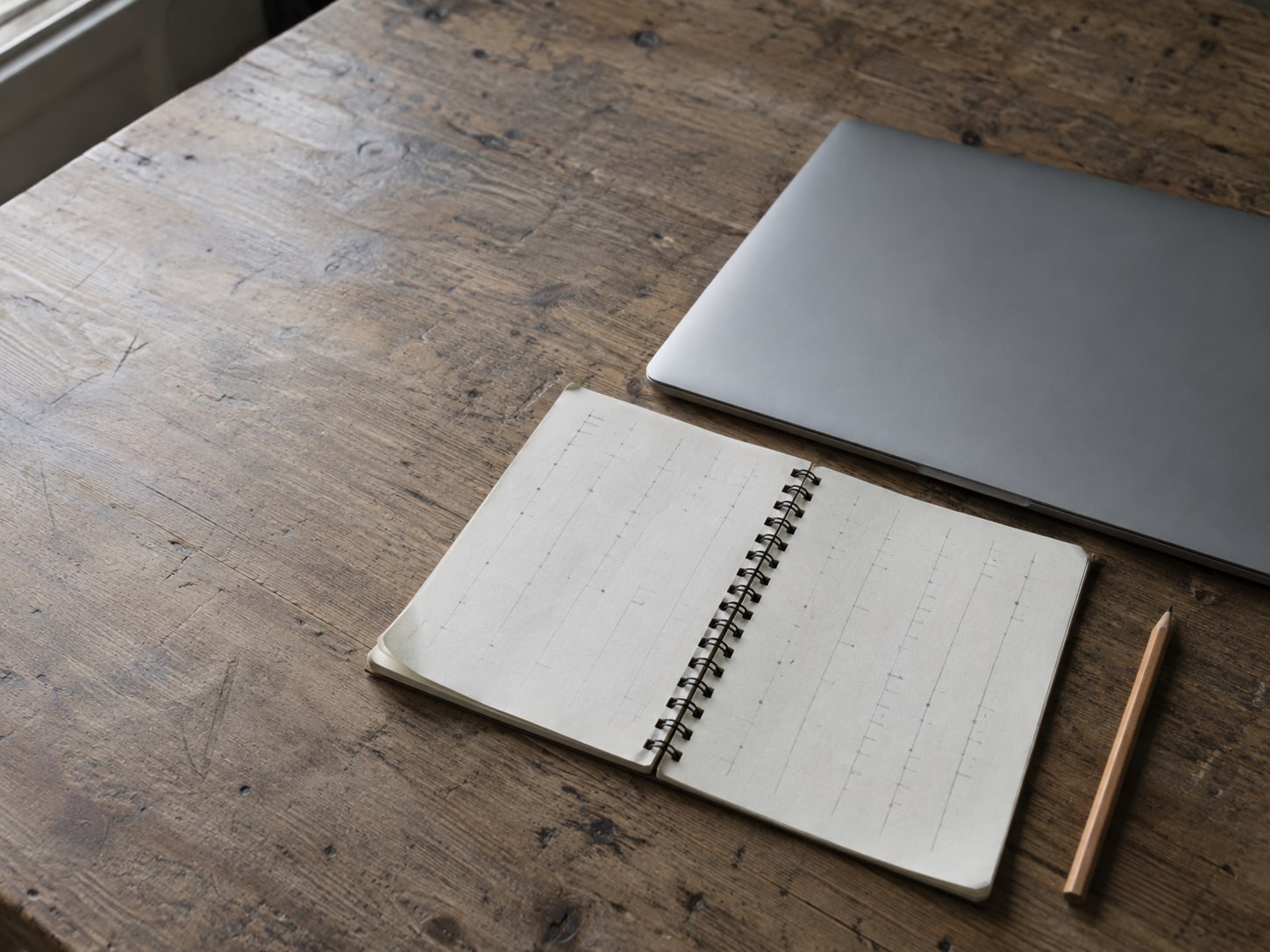 A spiral notebook lying open on a wooden desk with faint, unreadable pencil columns, next to a closed laptop and a sharpened pencil.