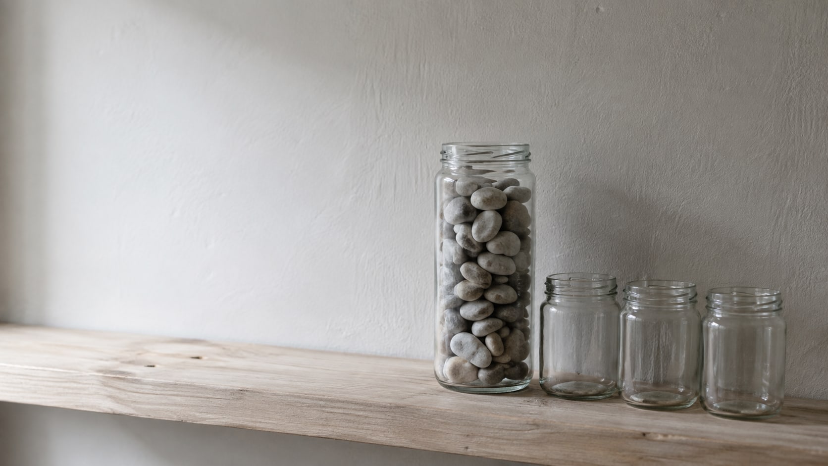 A tall glass jar full of smooth pebbles beside three nearly empty small jars on a pale wooden shelf in soft window light.