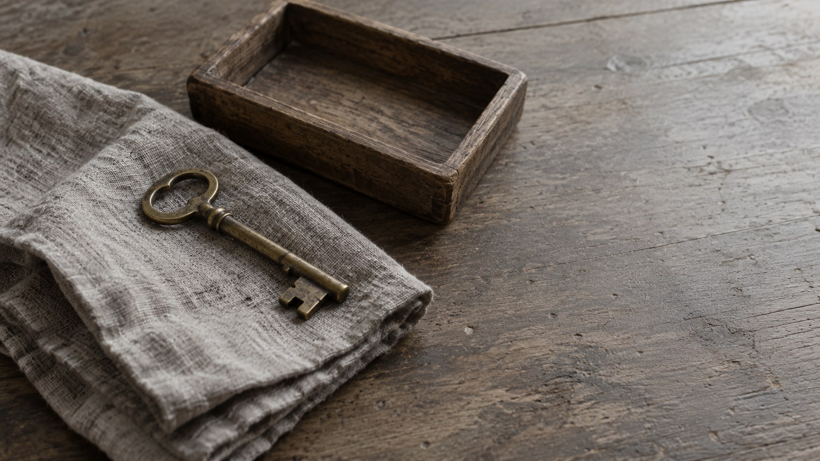 A single brass key resting on a folded gray cloth beside a small empty wooden tray on a plain tabletop.
