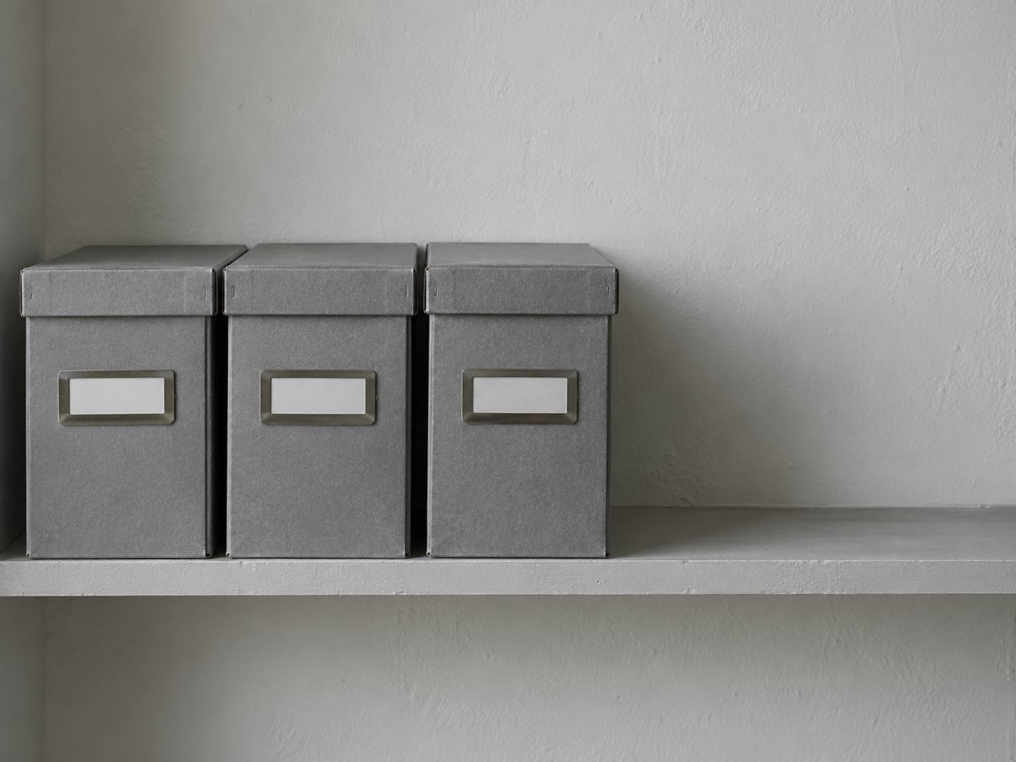 A tidy row of three identical gray filing boxes with blank labels on a shelf against a plain wall.