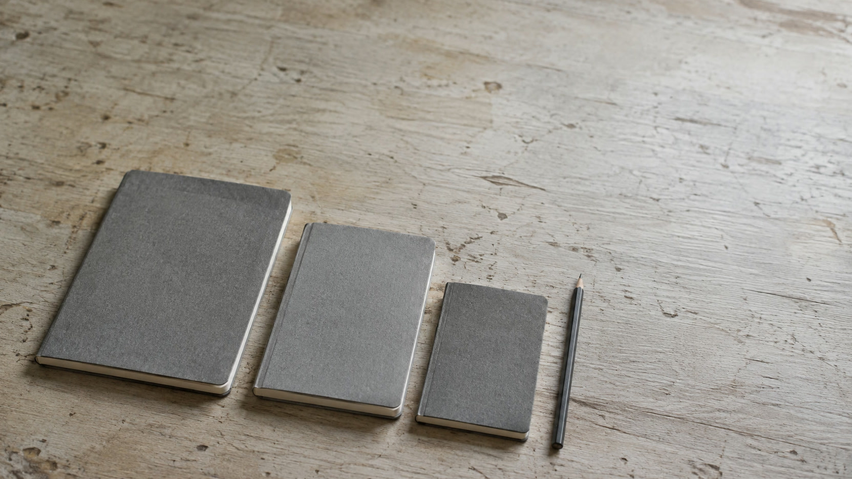 Three closed notebooks of different sizes lined up beside a pencil on a pale desk.