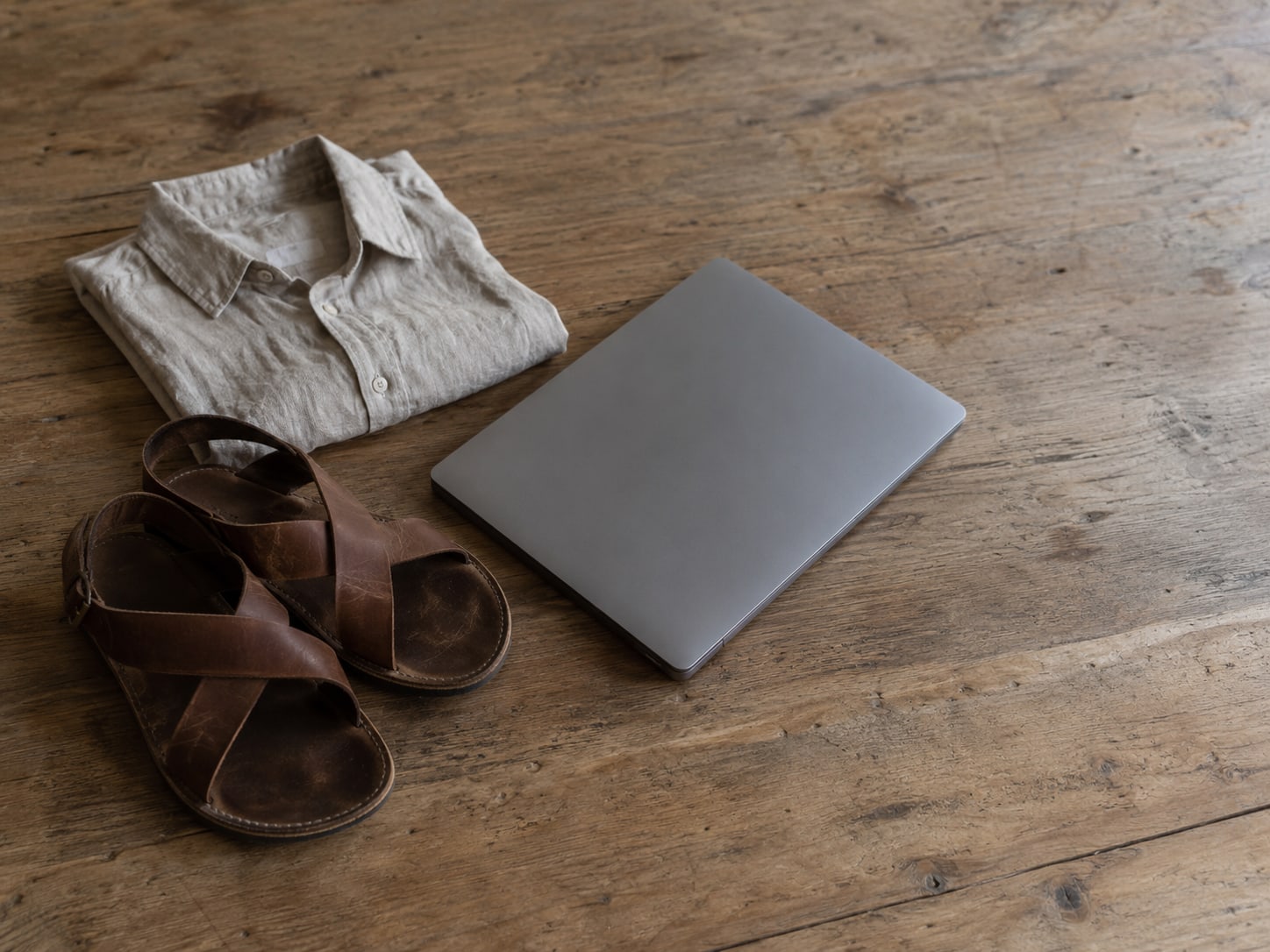 Una camisa de lino doblada y un par de sandalias de cuero sobre una mesa de madera lisa, junto a una laptop cerrada.