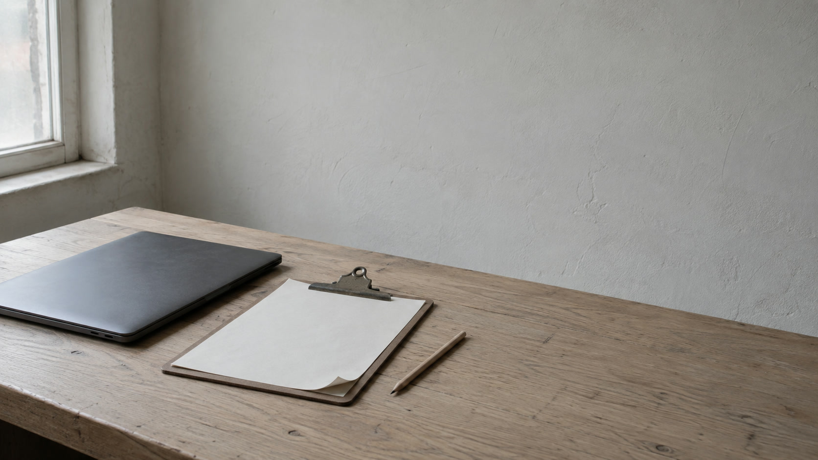 A closed laptop beside a clipboard holding blank paper and a pencil on a light wooden desk.