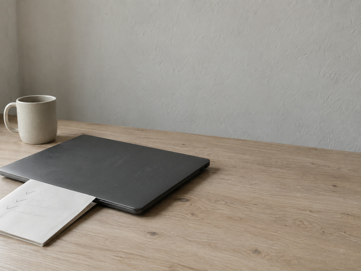 A closed laptop and a folded paper checklist with faint pencil ticks resting on a light oak desk beside a mug.