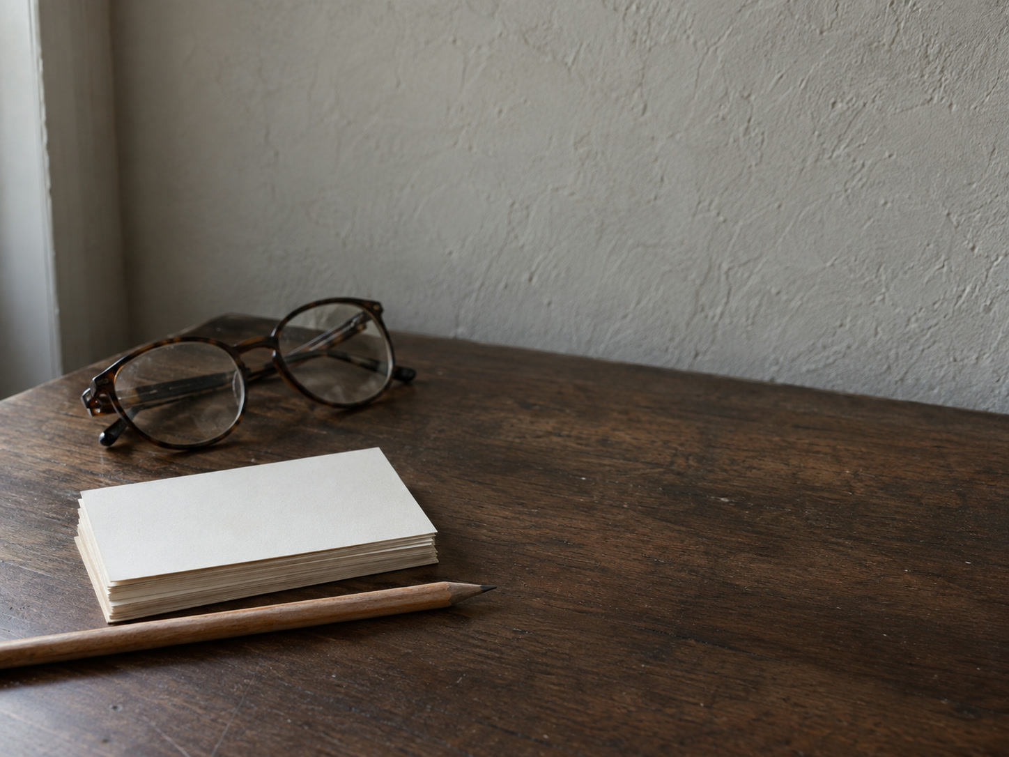 A stack of blank index cards beside a sharpened pencil and a pair of reading glasses on a dark walnut table.