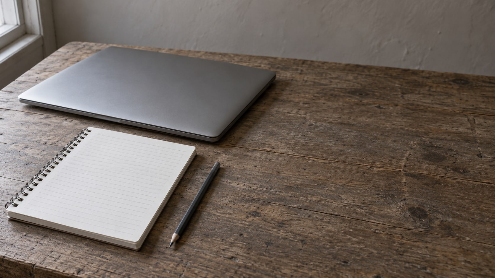 A closed laptop, a blank lined notepad and a pencil on a plain wooden desk.
