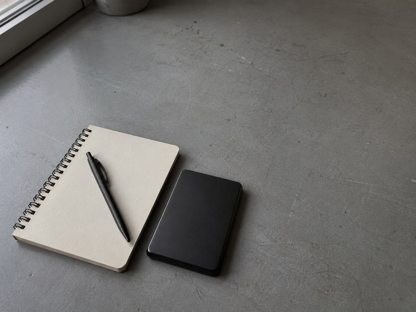 Un disco duro externo junto a un cuaderno de espiral cerrado con un bolígrafo encima, sobre un escritorio ordenado.