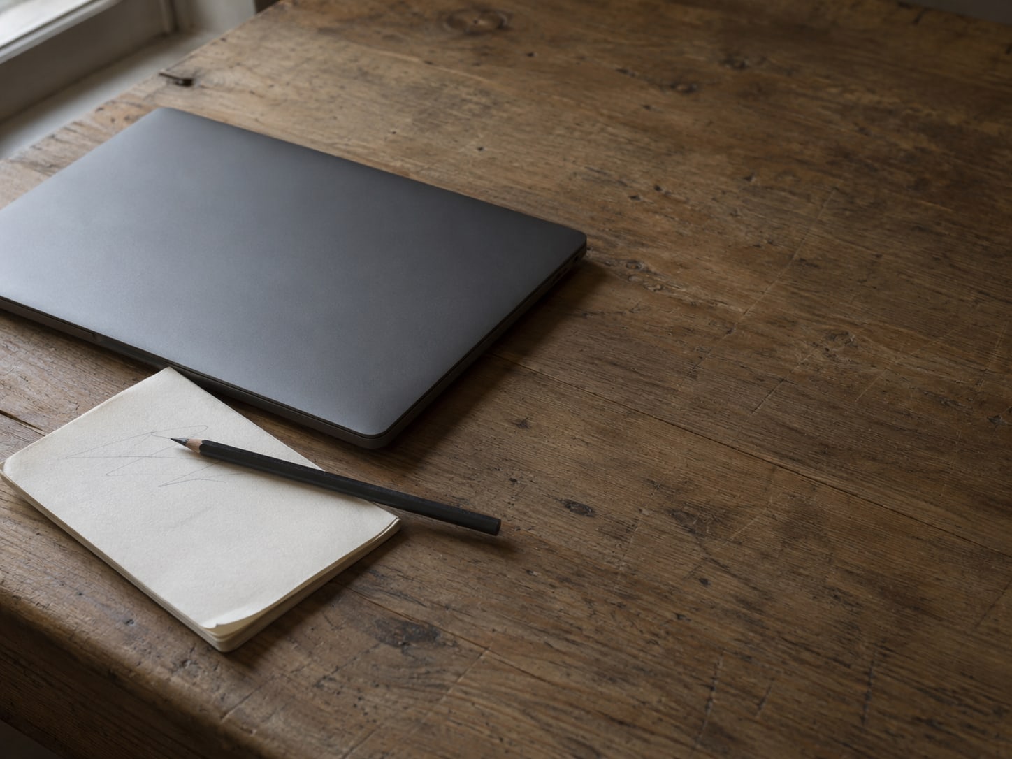 Una laptop cerrada junto a una pequeña libreta con marcas de lápiz y un lápiz atravesado sobre un escritorio de madera.