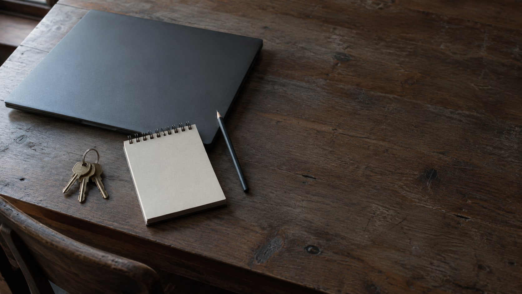 Una laptop cerrada y una pequeña libreta con un lápiz junto a un aro de llaves de latón sobre un escritorio de madera.
