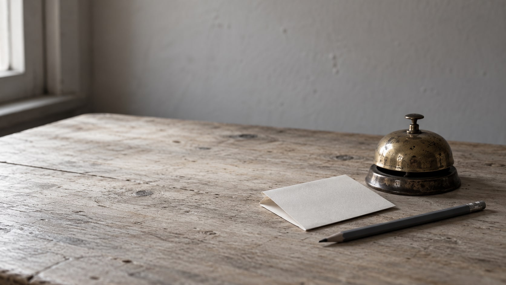 A small brass desk bell beside a folded paper note and a pencil on a light wooden desk.