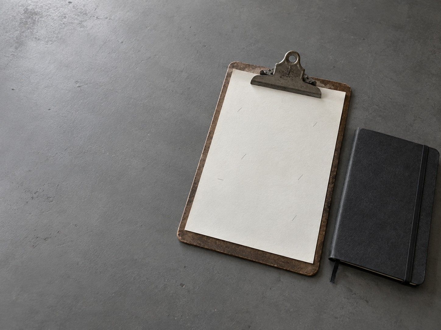 A clipboard holding a blank sheet with faint pencil tick marks beside a closed notebook on a gray desk.