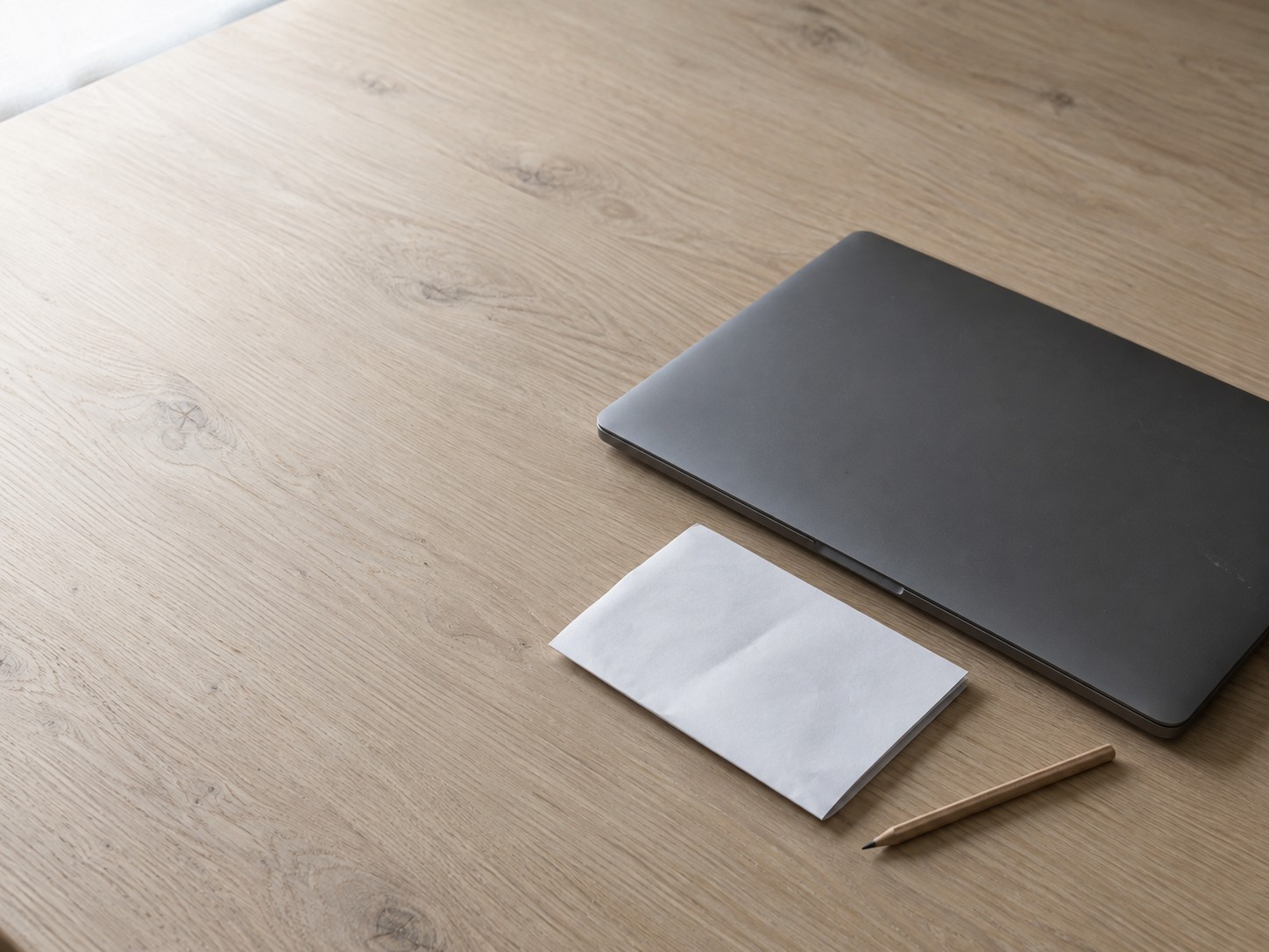 A closed laptop beside a folded paper billing statement and a pencil on a plain oak desk.