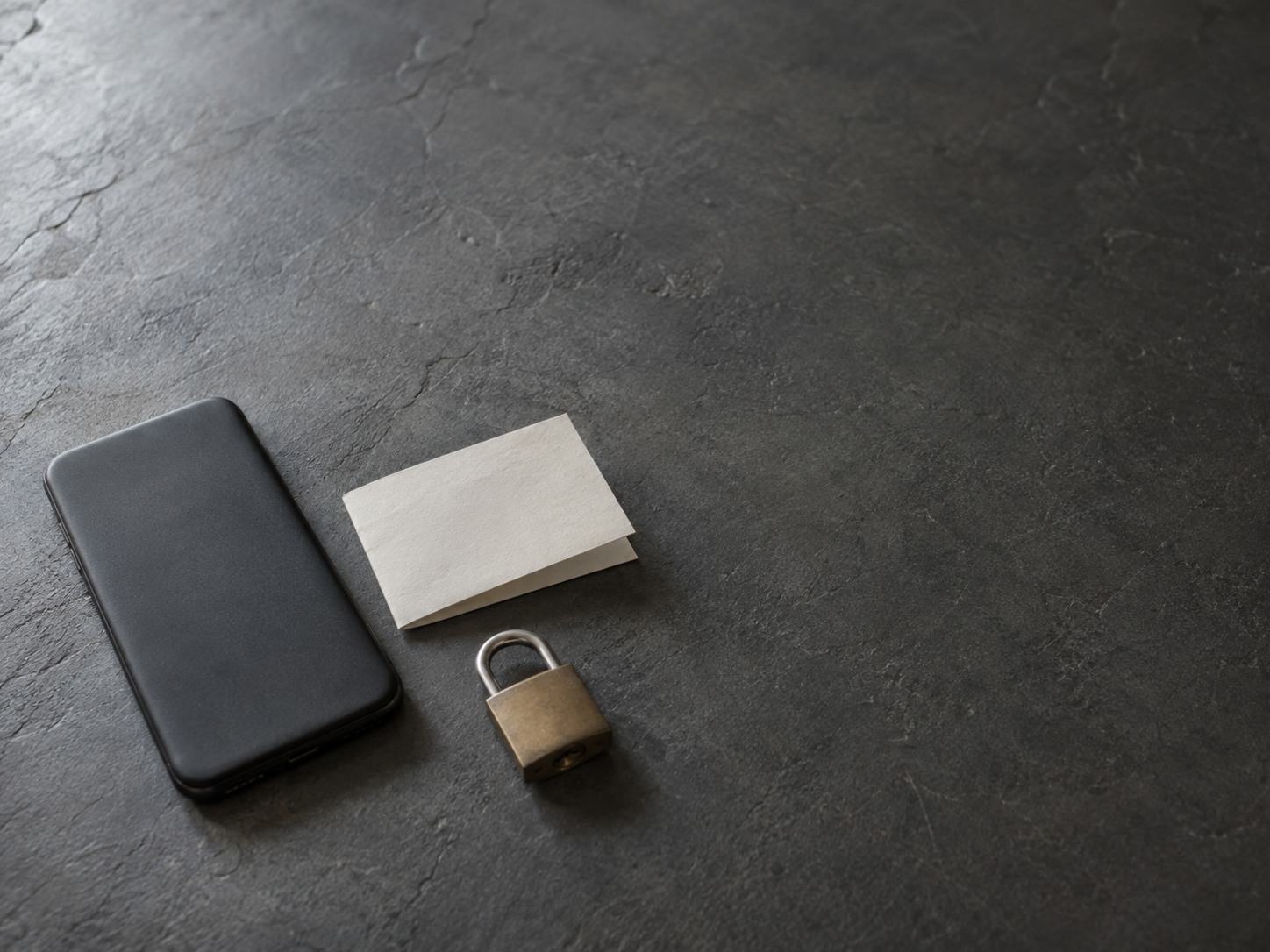 A switched-off phone lying face down beside a folded blank note and a small closed padlock on a slate surface.