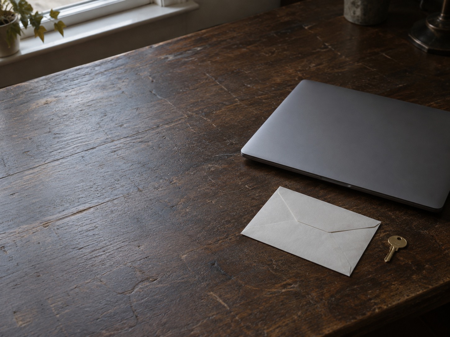 A closed laptop beside an unopened envelope and a small brass key on a worn wooden desk.