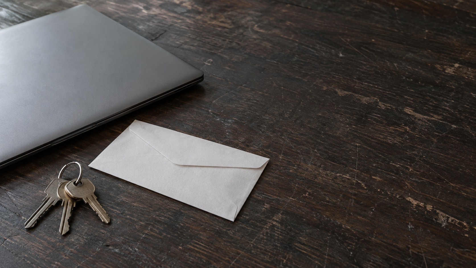 A ring of keys beside an unopened envelope and a closed laptop on a dark desk.