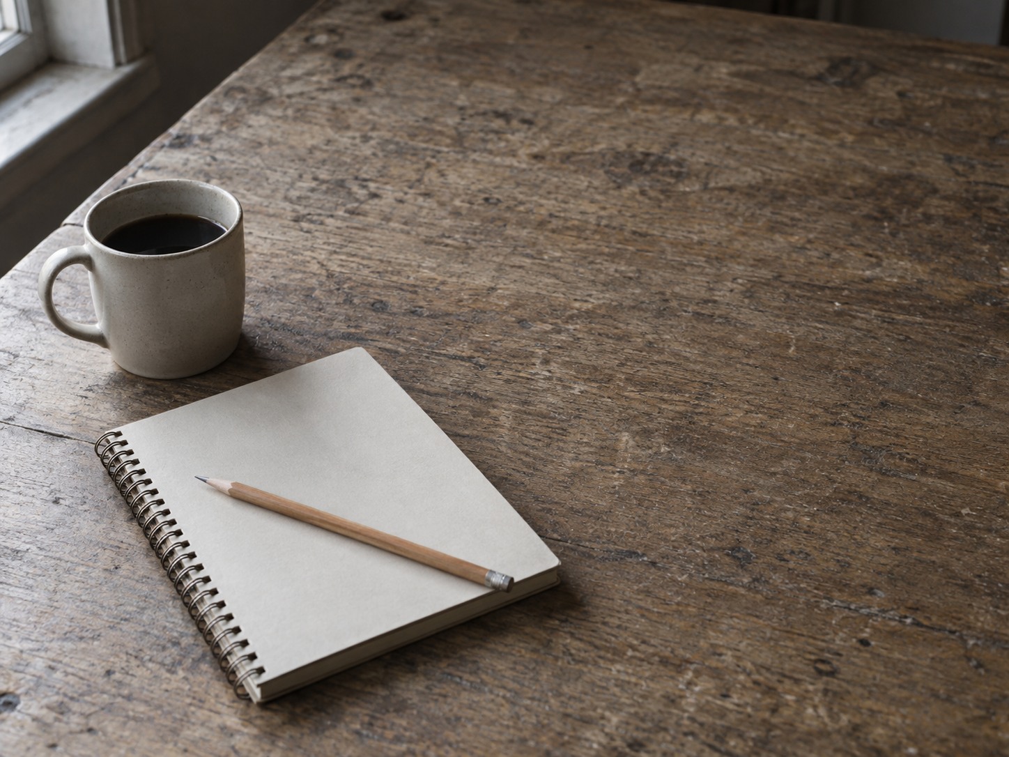 A closed spiral notebook with a pencil resting across it next to a coffee mug on a plain wooden desk.