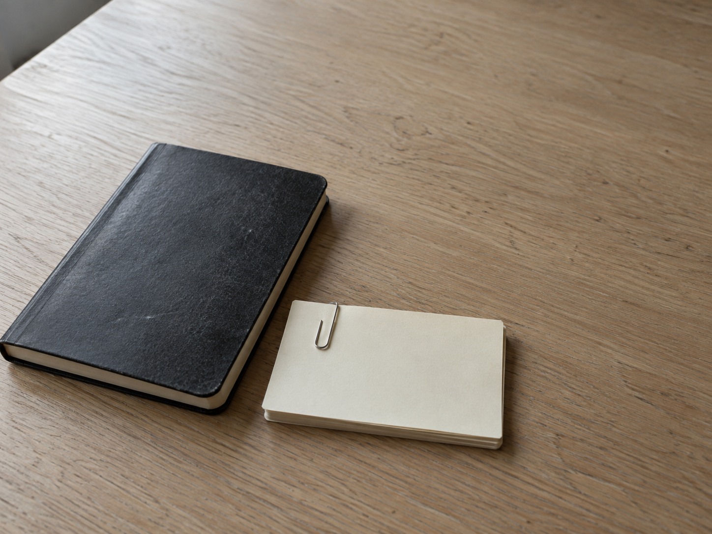 A small stack of blank index cards held with a paper clip next to a closed notebook on a light oak table.