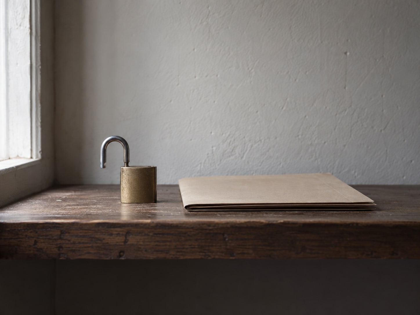 A padlock resting open on a wooden shelf next to a neatly closed folder, lit by soft window light.