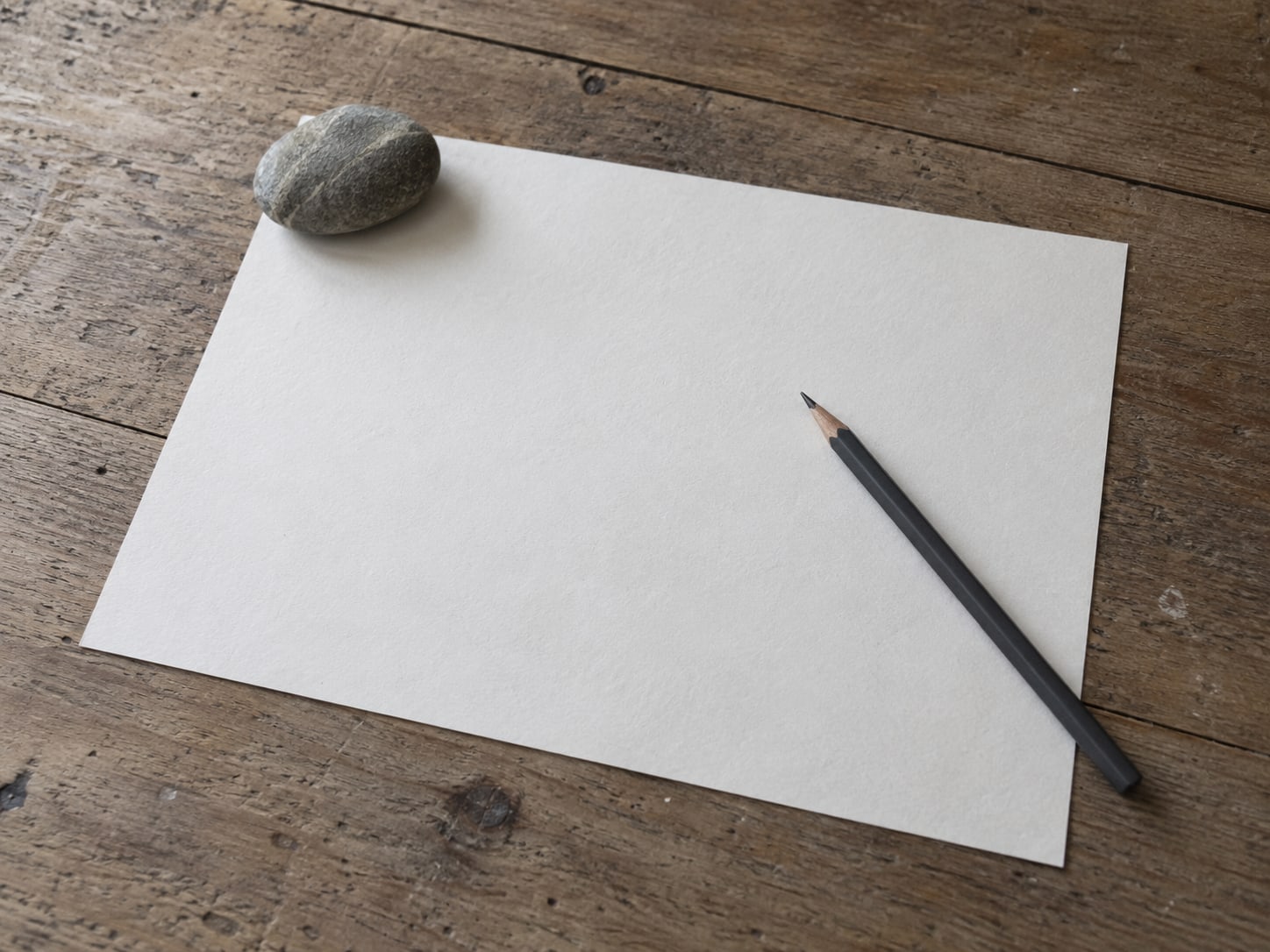 A paper calendar page held flat by a smooth stone, with a pencil resting on top.