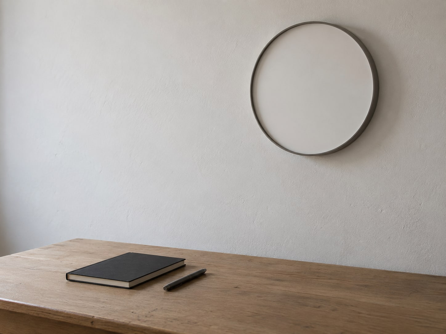 A wall clock with a plain face above a tidy desk holding a closed notebook and a pen.