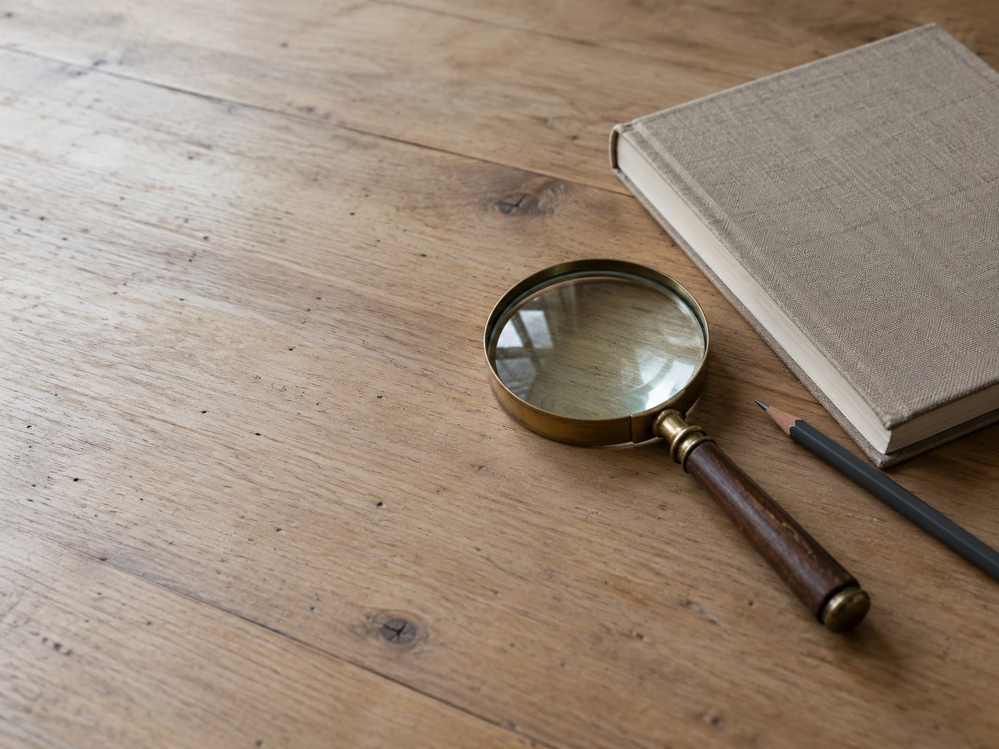 A magnifying glass resting on a plain wooden desk beside an unopened notebook and a pencil.