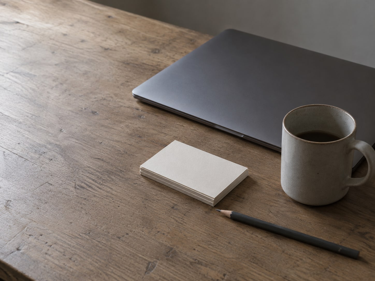 A closed laptop, a small stack of blank index cards and a pencil resting on a wooden desk beside a mug.