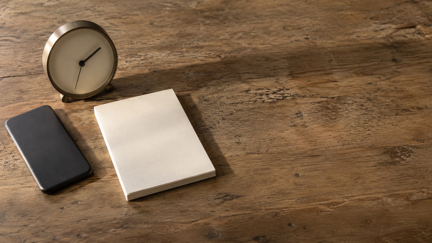 A closed smartphone beside a small desk clock and a blank notepad on a wooden desk in evening light.