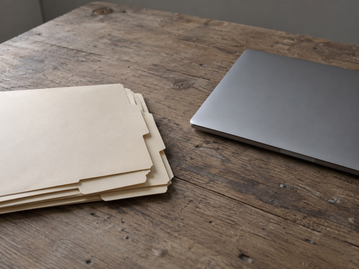 A neat stack of paper folders with blank tabs beside a closed laptop on a wooden table.