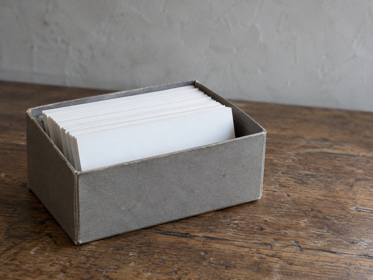 A gray archive box with its lid off, holding a row of blank index cards, on a wooden table.