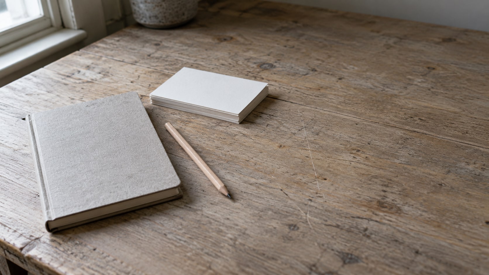 A closed notebook, a pencil and a small stack of blank index cards arranged on a wooden desk.