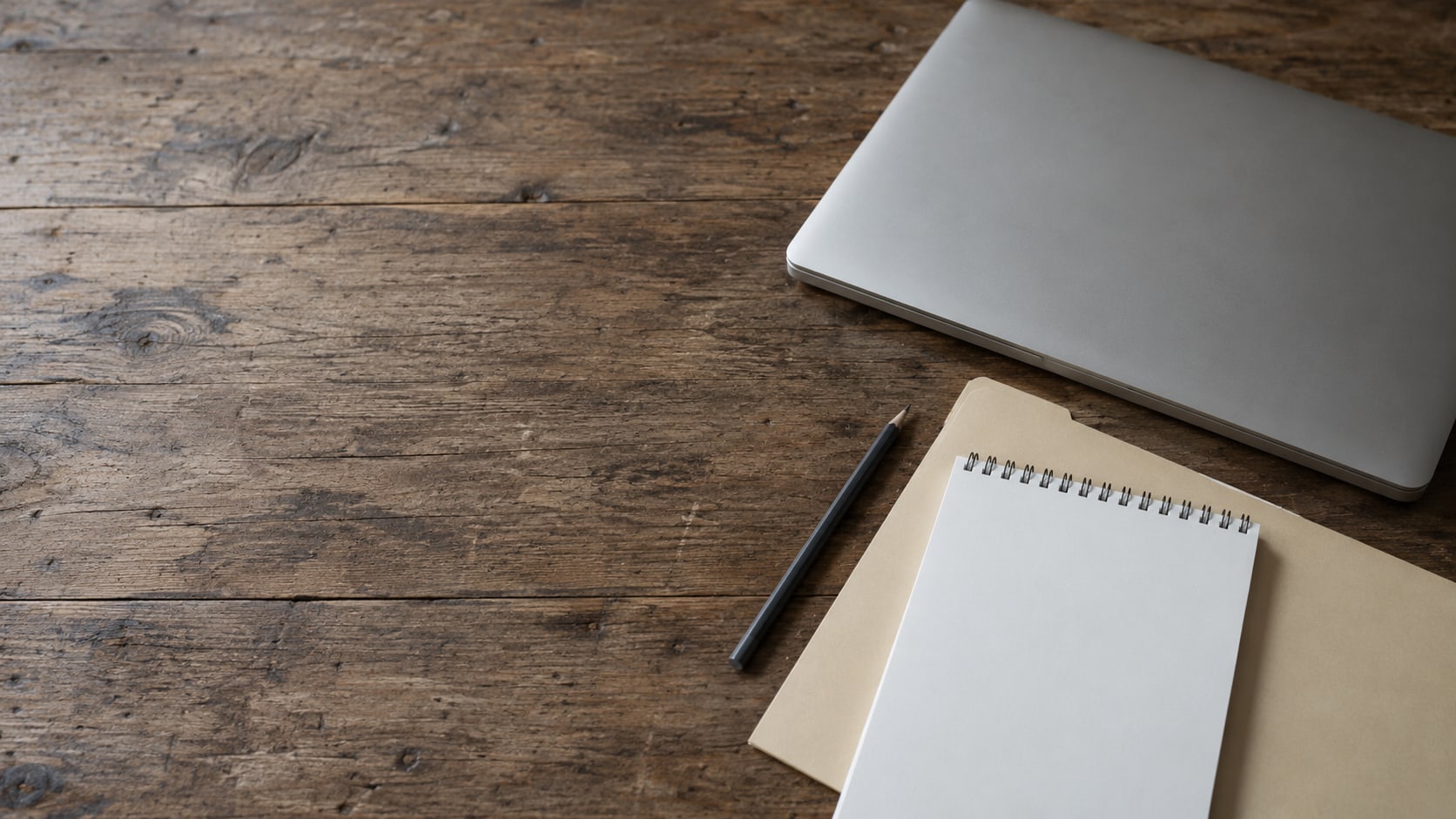 A closed laptop, a manila folder and a pencil resting on a worn wooden desk beside a blank notepad.