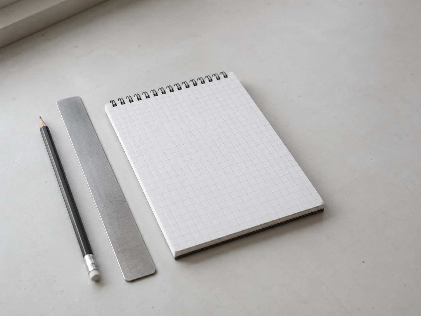 A pencil, a ruler and a blank grid-lined notepad lined up on a pale desk surface.