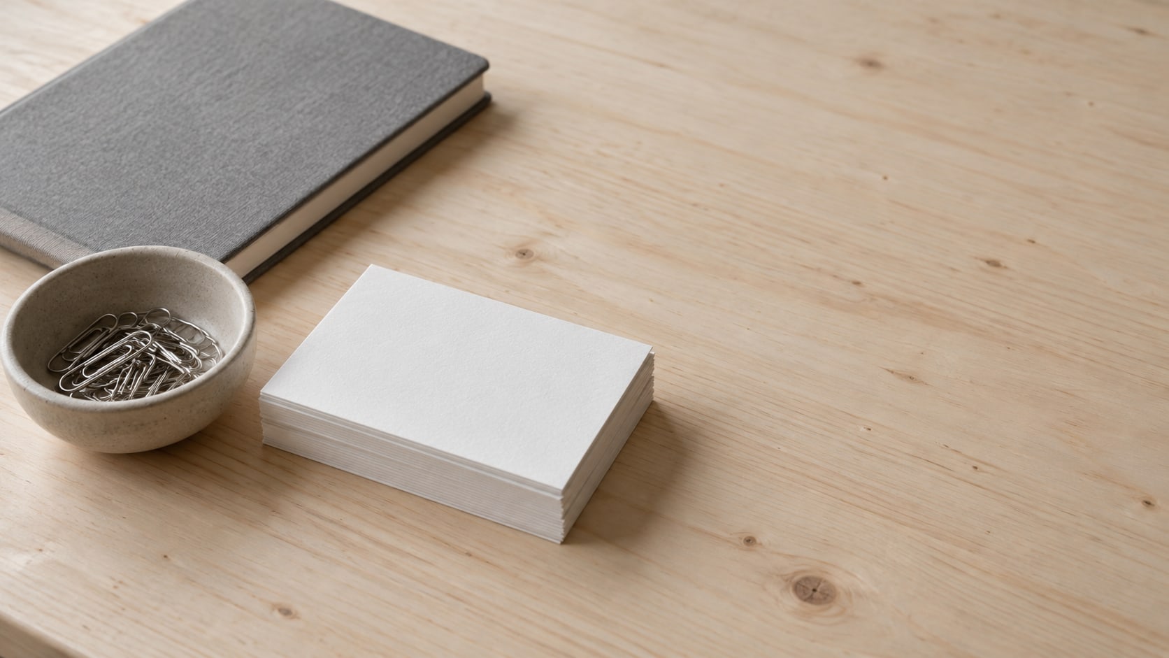 A stack of plain blank cards and a small ceramic bowl of paperclips on a light wooden counter, with a closed notebook beside them.