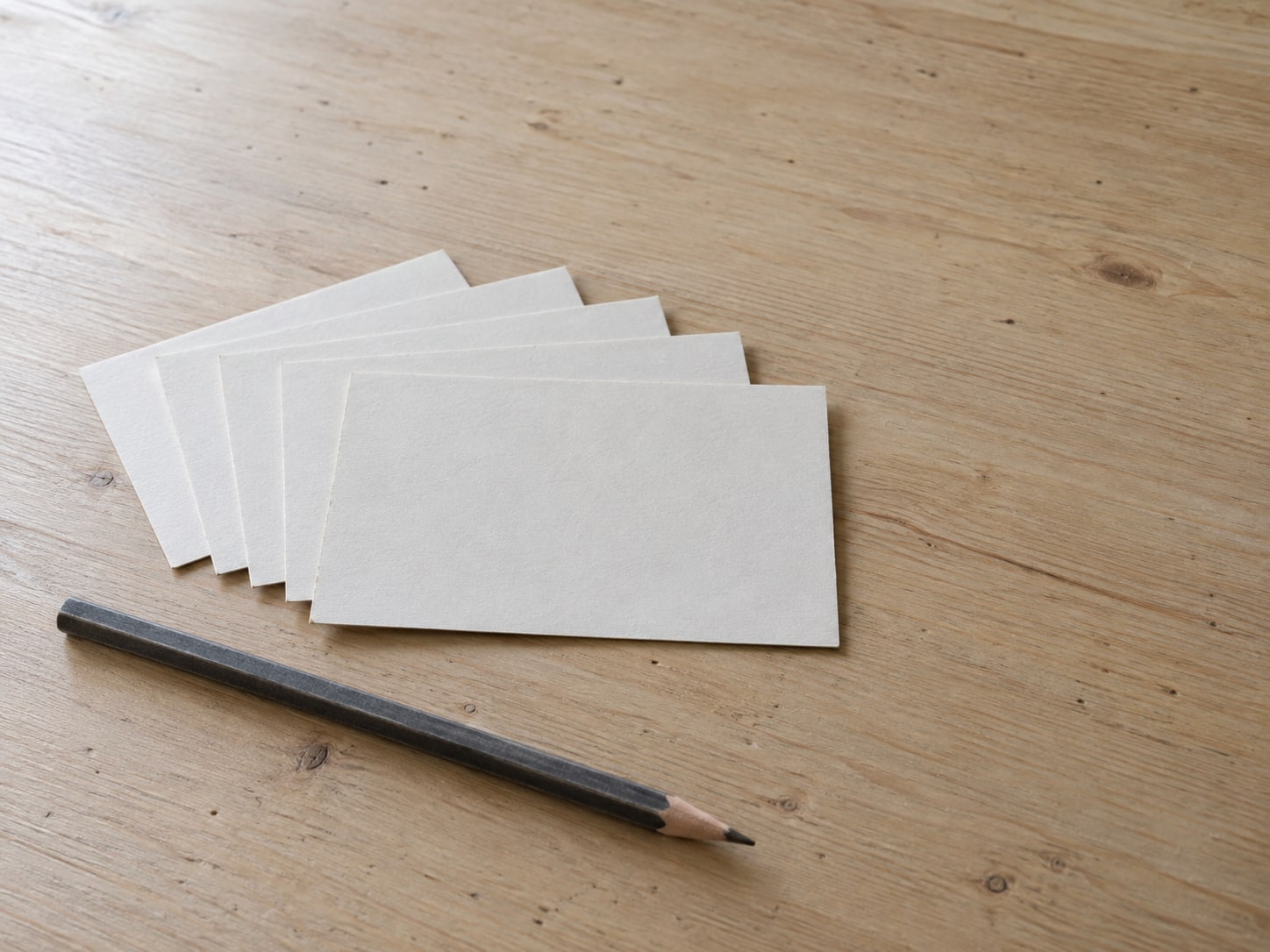 Five blank index cards fanned out beside a pencil on a plain wooden desk.
