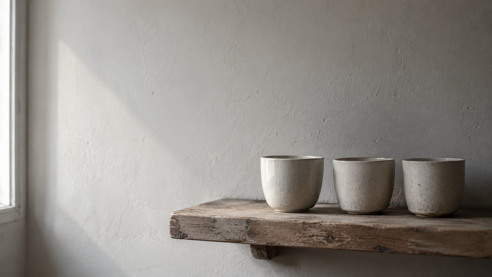 Una fila de tres tazas de cerámica sobre una repisa de madera sin pintar, una de ellas girada hacia la luz suave de una ventana.